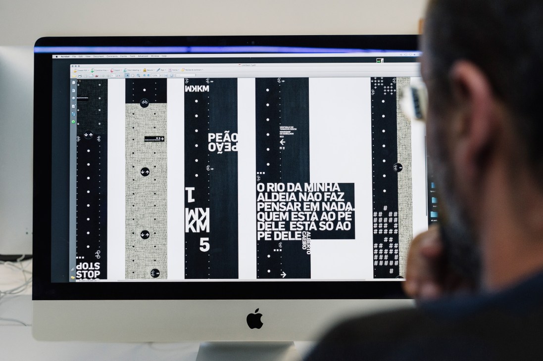Designer working on computer displaying cycle lane layout designs with black and white graphics and Portuguese text.