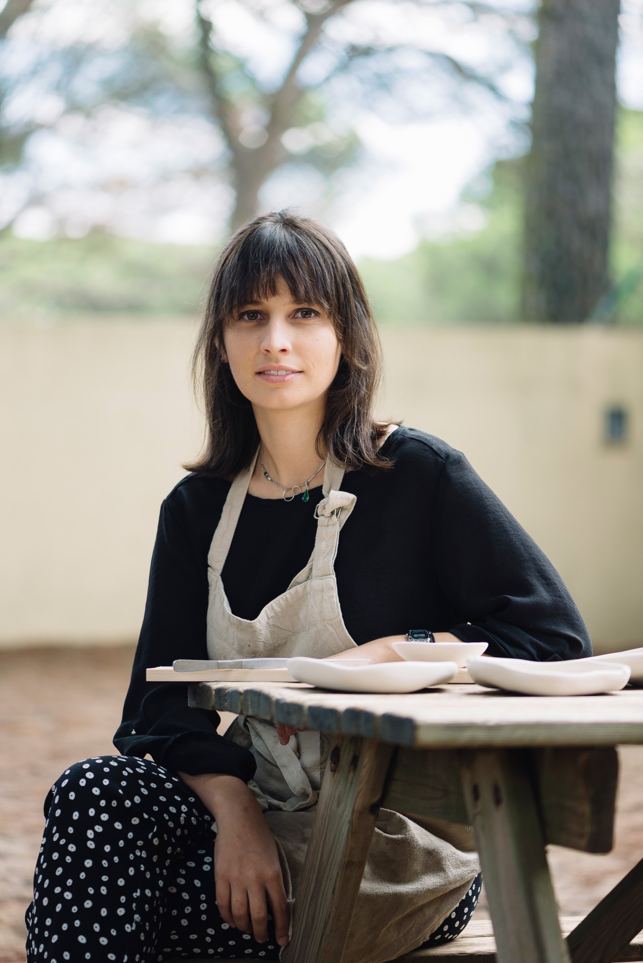 Margarida Almeida, a young ceramic designer, sits outdoors wearing an apron with ceramic pieces displayed before her.