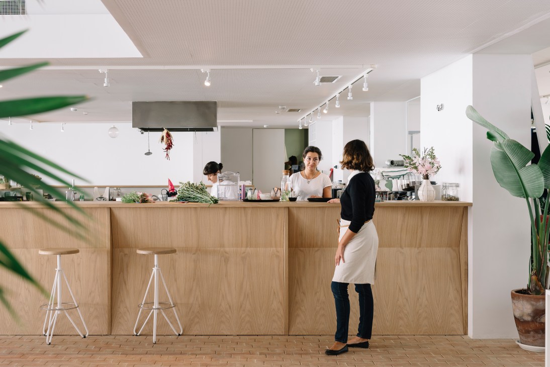 Woman standing at wooden hotel bar counter with staff behind, white stools, plants and track lighting.