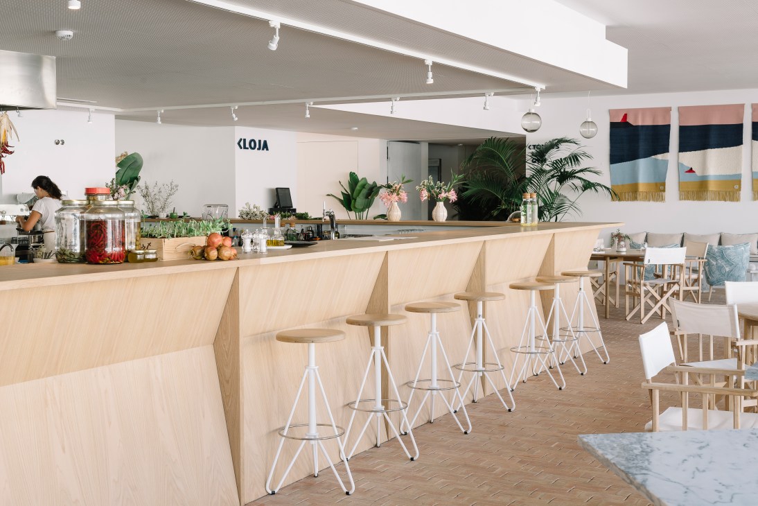 Modern hotel bar with wooden counter, white bar stools, and plants, designed by Martin Blanchard.