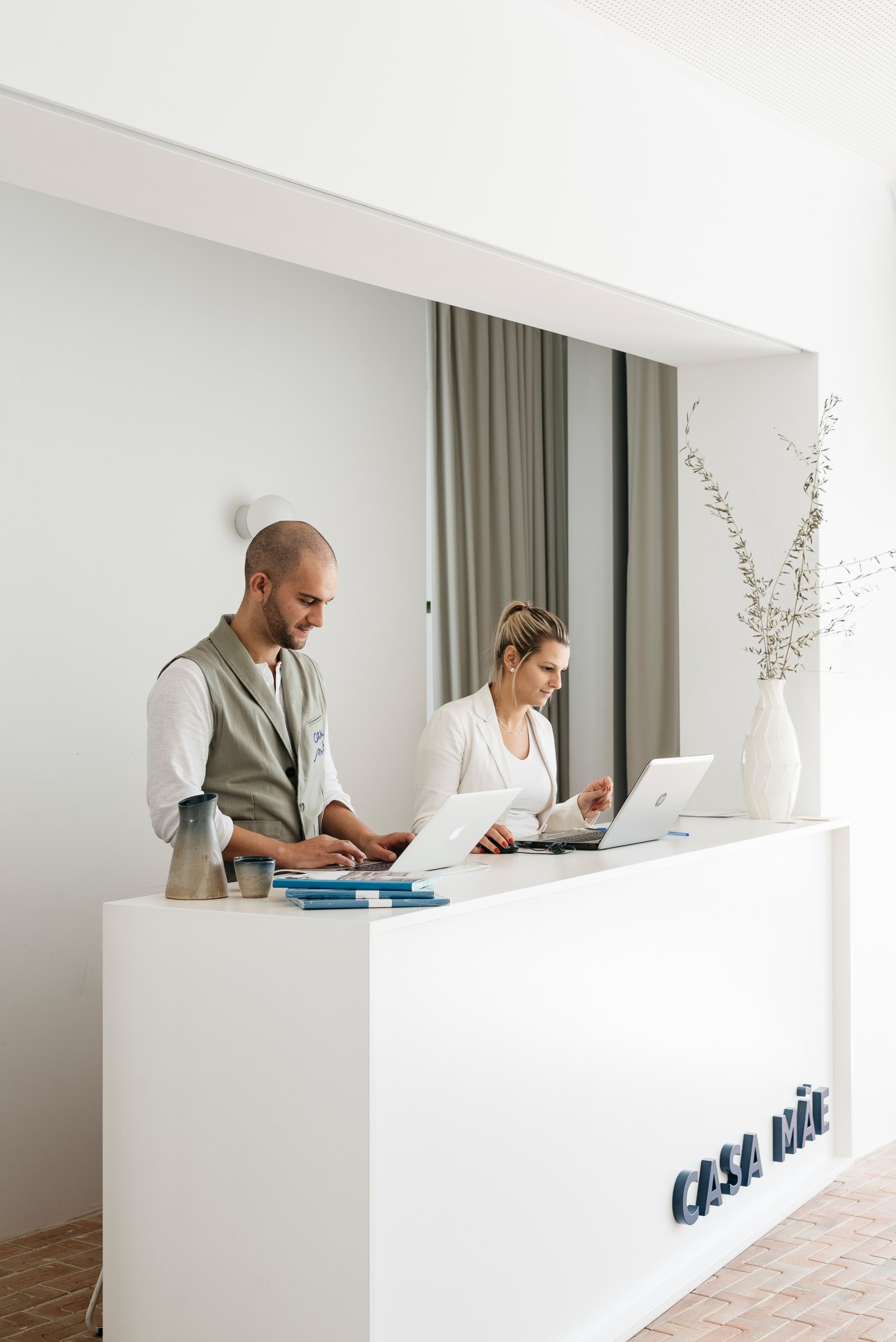 Two hotel staff members at Casa Mãe's white reception desk, one wearing a vest designed by La Paz.