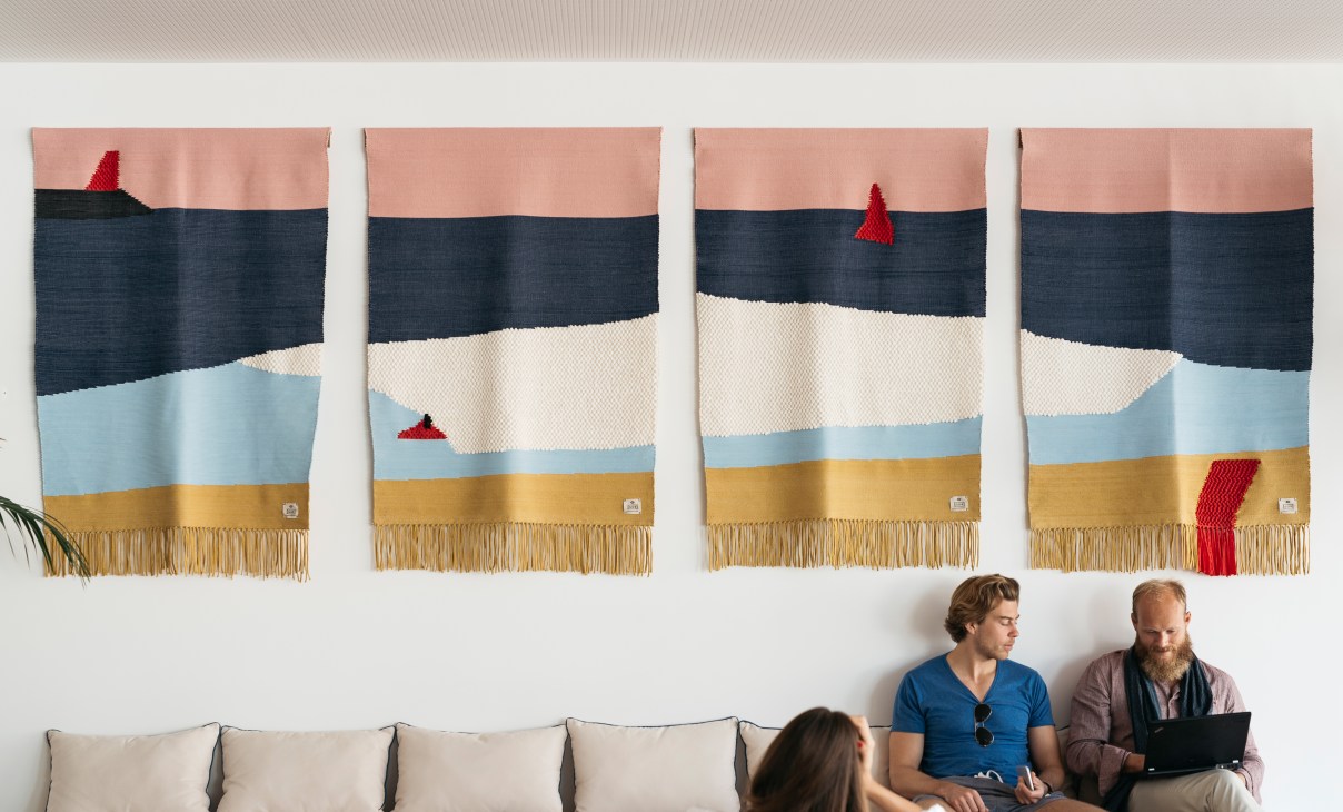 Four colorful woven rugs with horizontal stripes in pink, navy, cream, blue and gold with red tassels hang on a white wall above people seated on a bench.