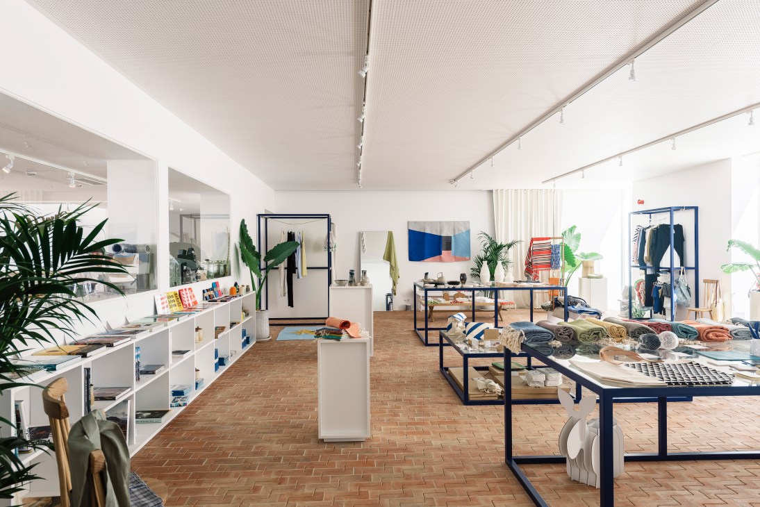 Bright retail shop interior with white shelving, terracotta brick floors, and displayed clothing and design products.