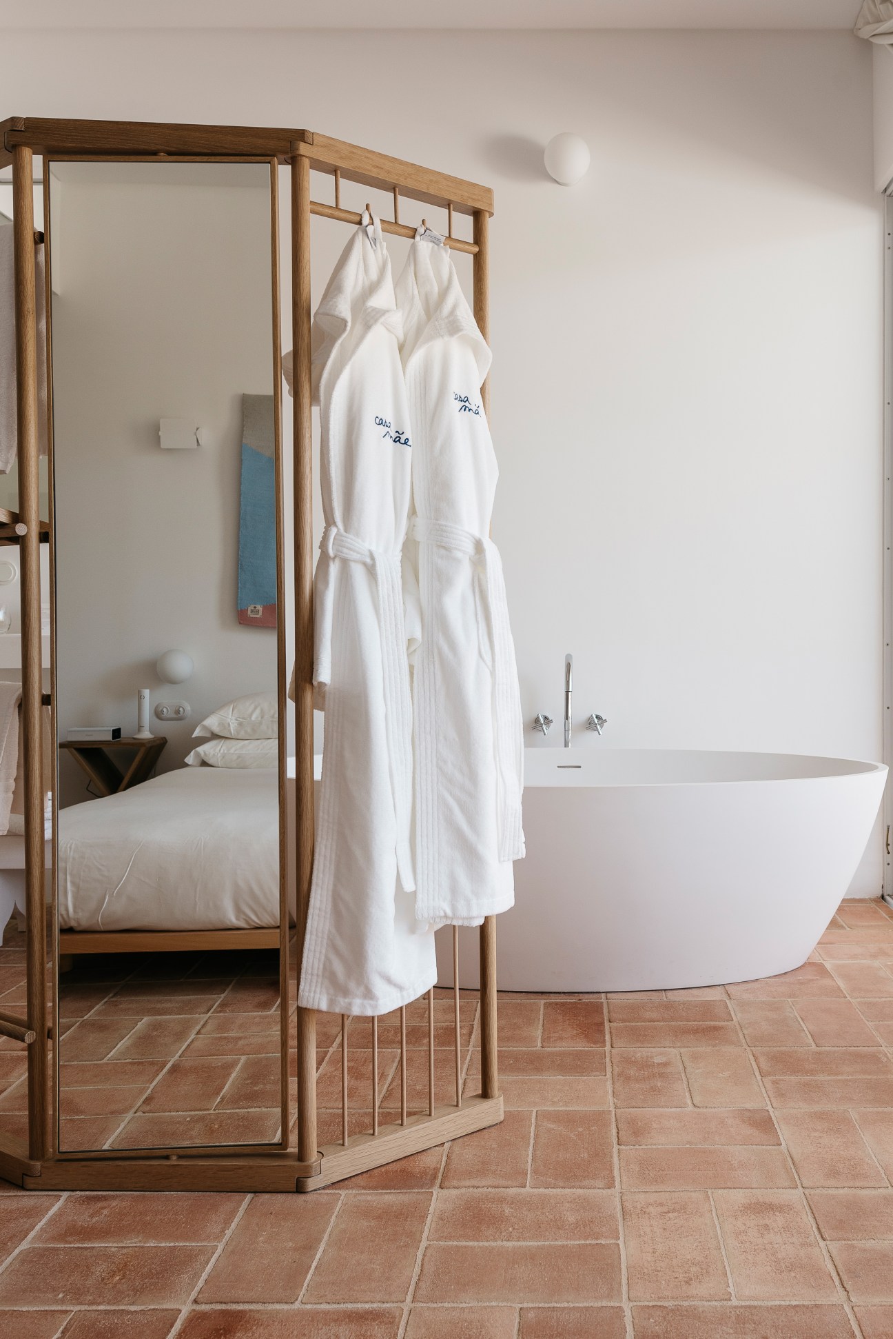 Wooden-framed mirror screen with white robes hanging on rod, separating bedroom from modern white freestanding bathtub on terracotta tiles.