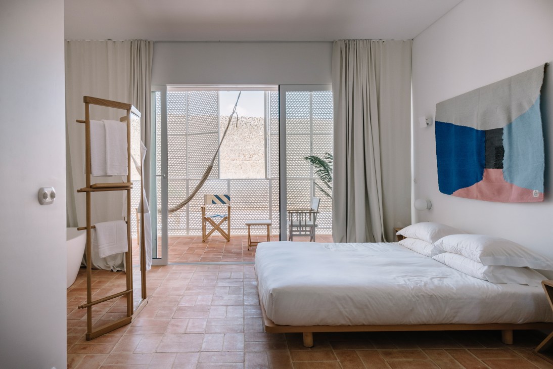 Bright modern hotel bedroom with terracotta floors, wooden bed, oak screen divider, and balcony with geometric lattice screens overlooking the Algarve landscape.