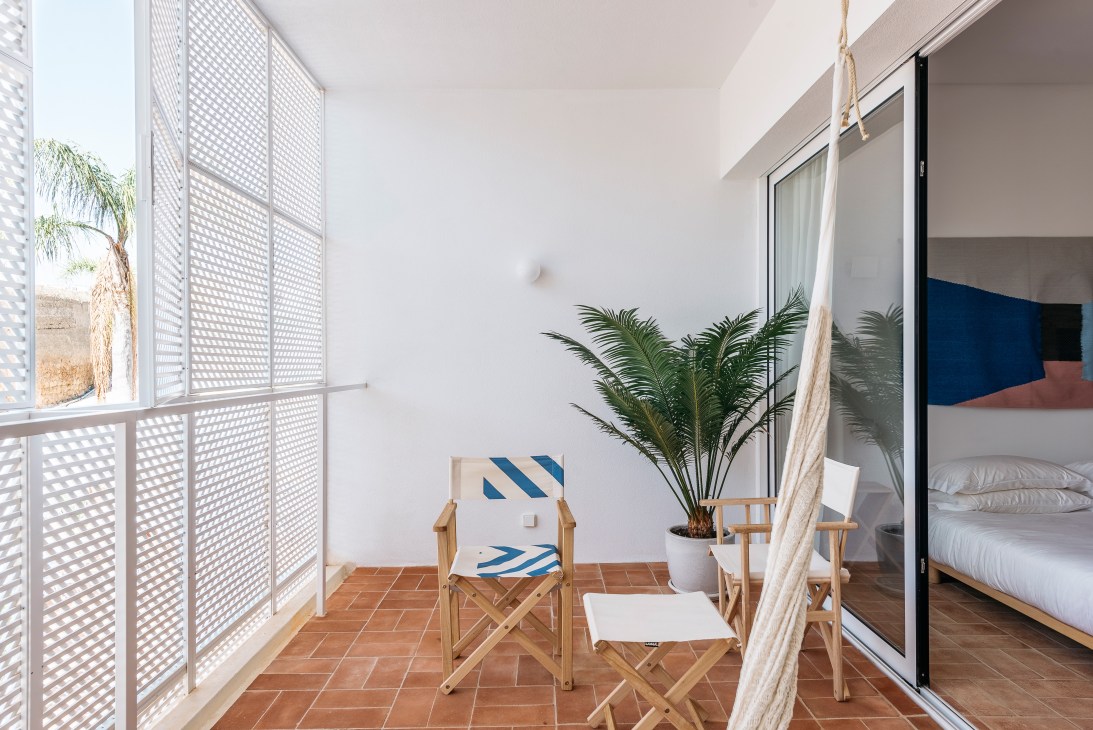 A bright bedroom balcony with wooden deck chairs, potted palm plant, terracotta tiles, white walls, and geometric wooden screens.