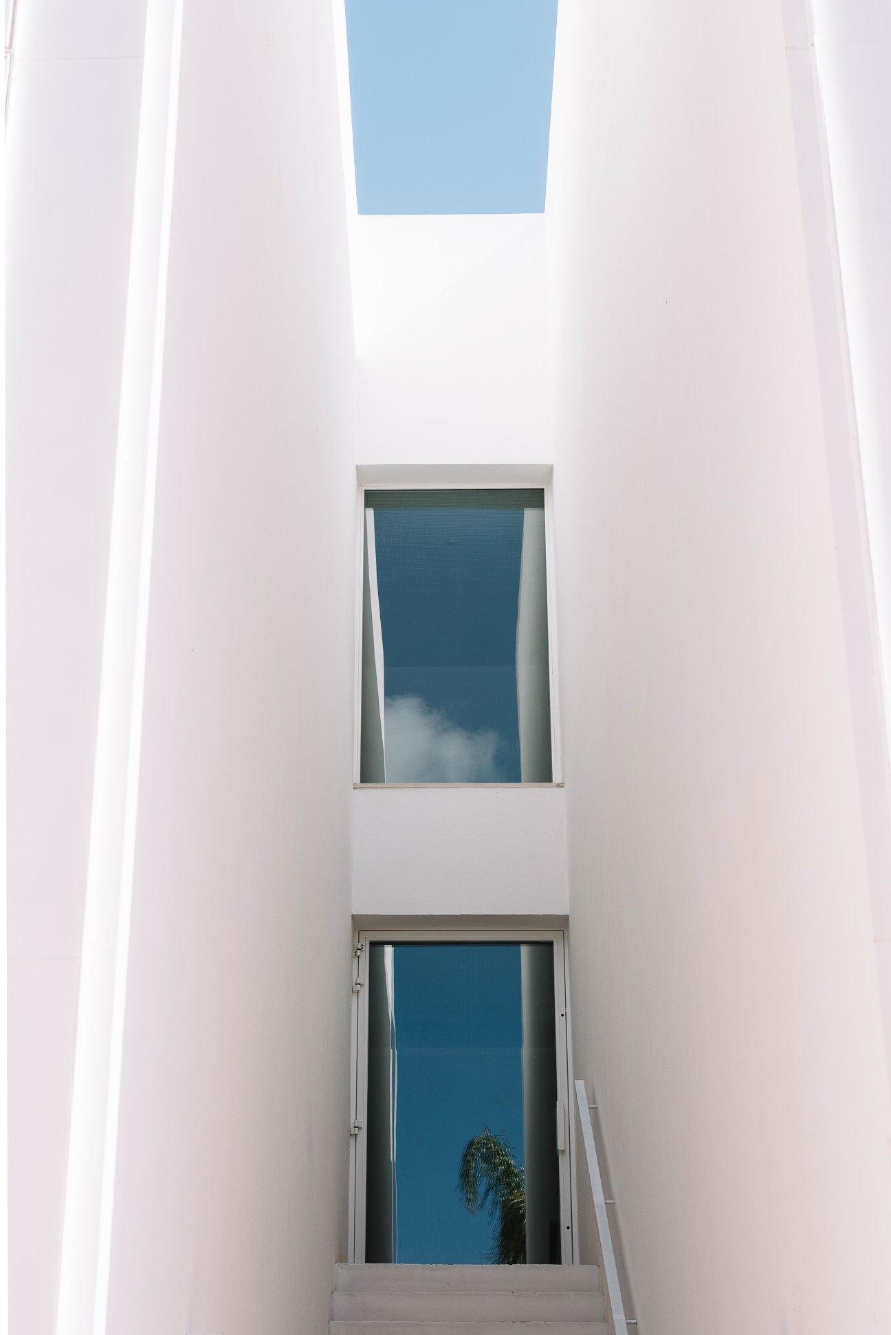 White-walled hotel exterior with geometric windows framing blue sky and a palm plant.