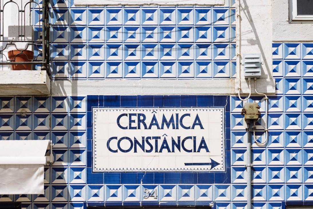 White ceramic sign with blue text reading 'CERAMICA CONSTÂNCIA' and an arrow, mounted on blue and white geometric patterned tiles.