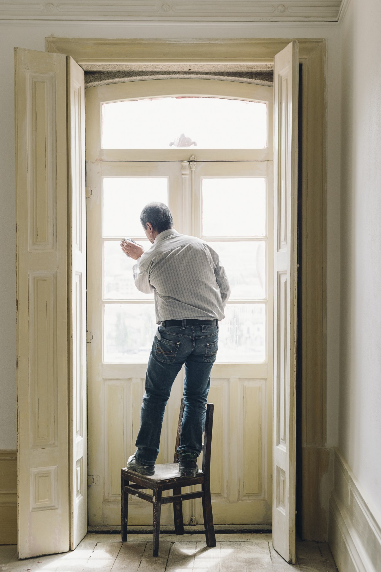 Worker standing on chair at tall window with cream-colored frame during building renovation.