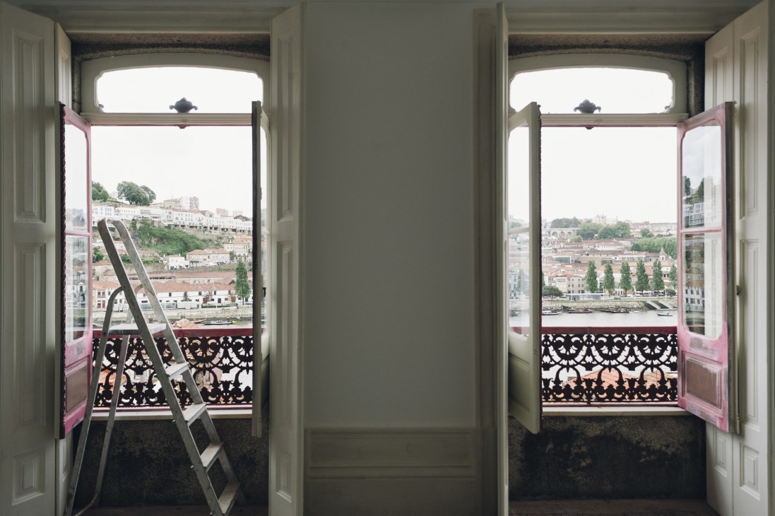 Open doors with ladder frame the River Douro and Porto cityscape beyond ornate balcony railing.