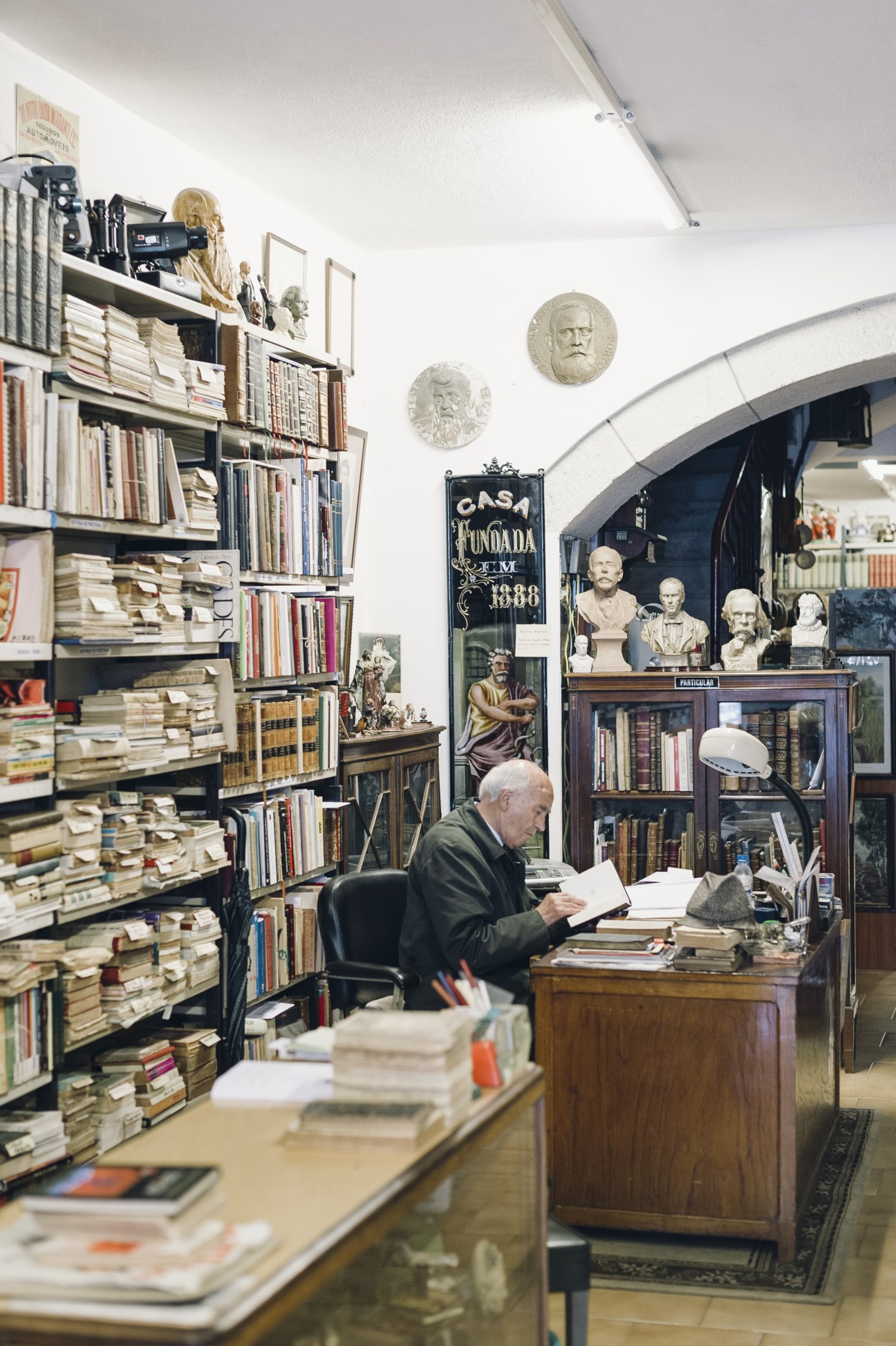 An elderly man browses books at a wooden counter in a vintage bookshop with shelves and busts.