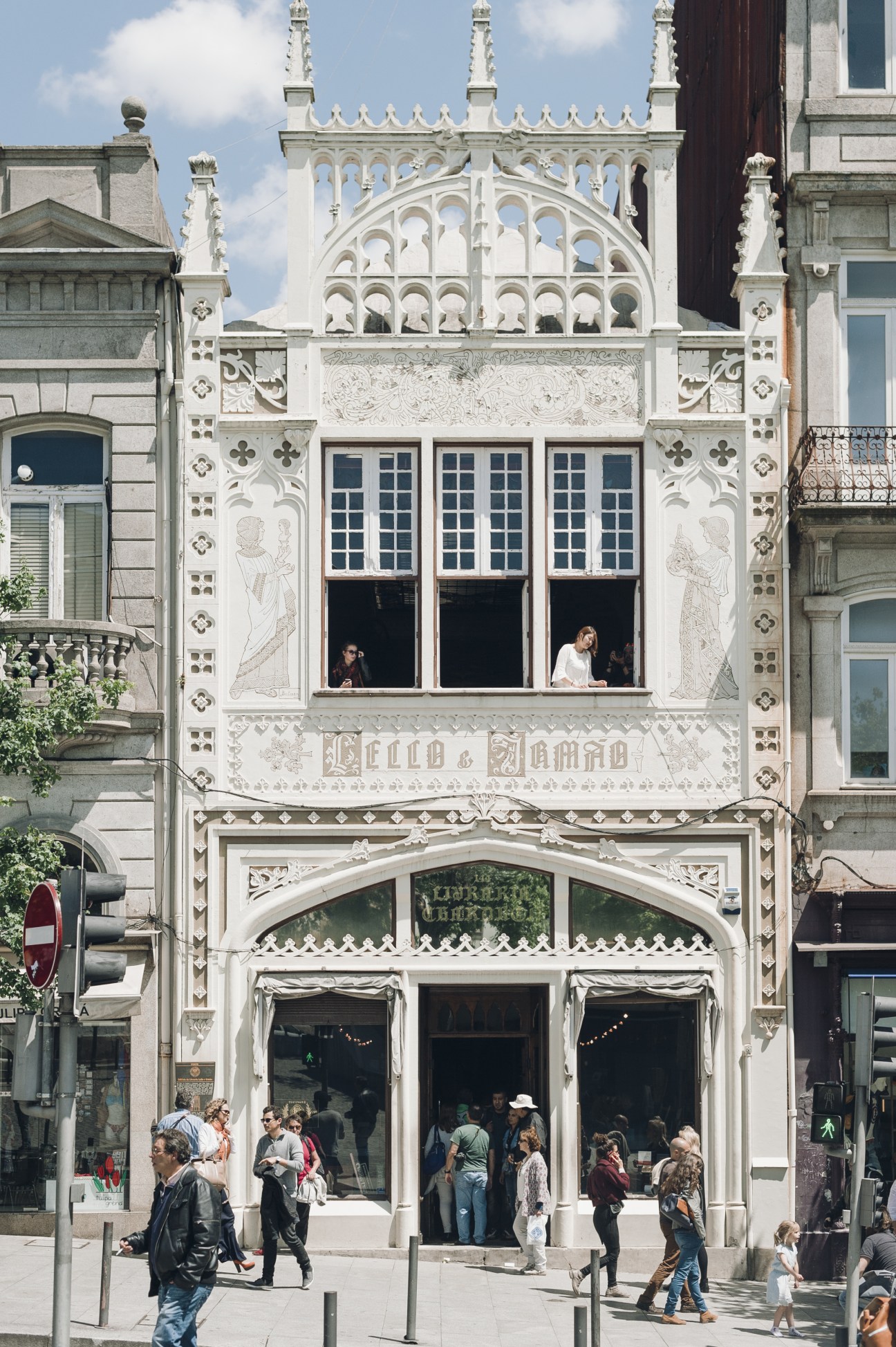Ornate white historic building facade with decorative stone carvings and large windows, featuring a ground-floor shop with pedestrians passing by.