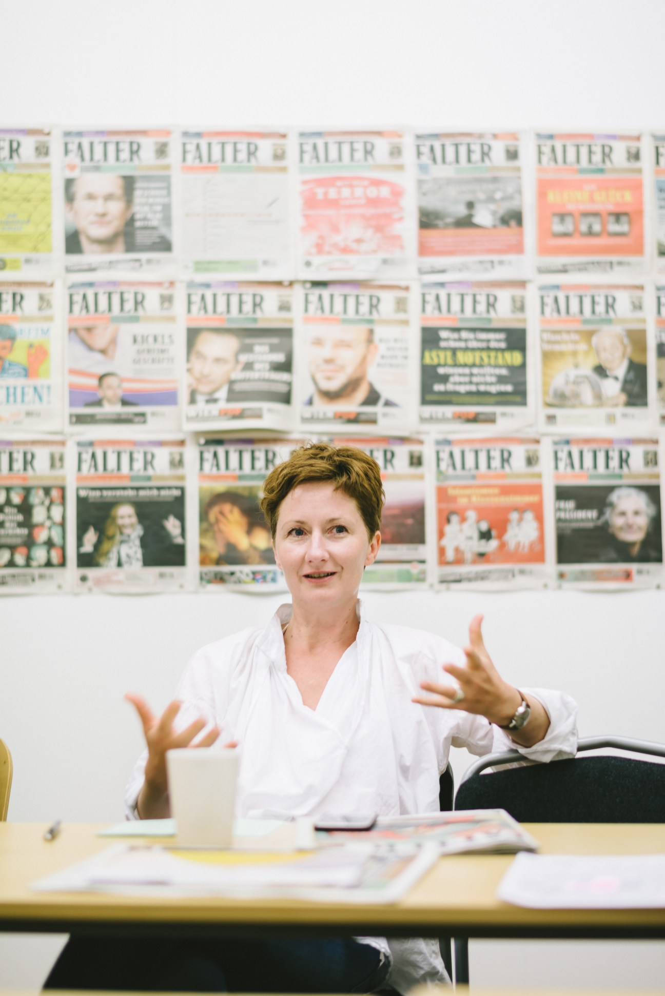 Editor Barbara Tóth gestures while speaking at a desk, with Falter magazine covers displayed on the wall behind her.