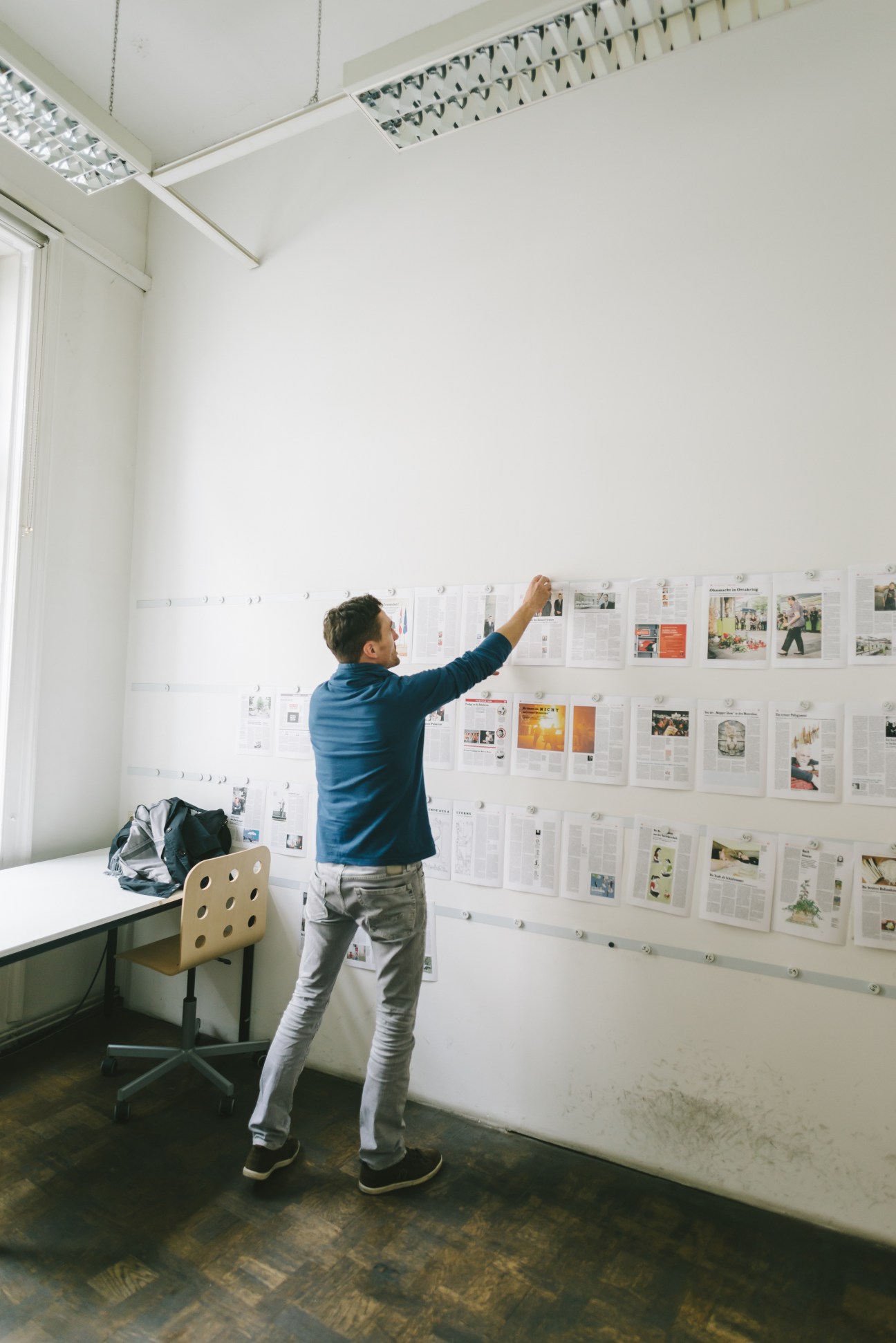 Man in blue jacket arranging newspaper layouts on white wall in bright office