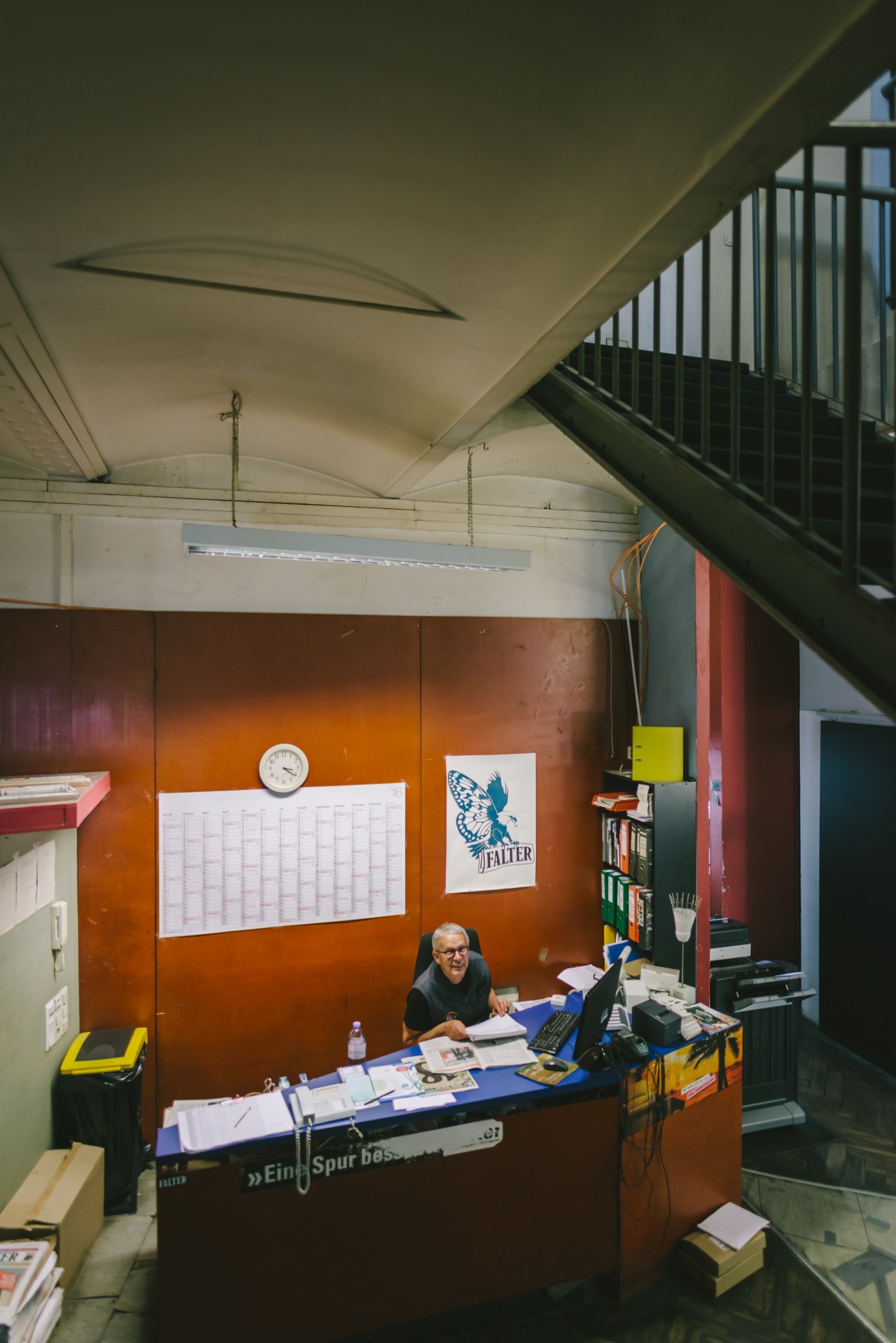 Person working at front desk in Falter magazine headquarters with orange wall and butterfly logo visible.