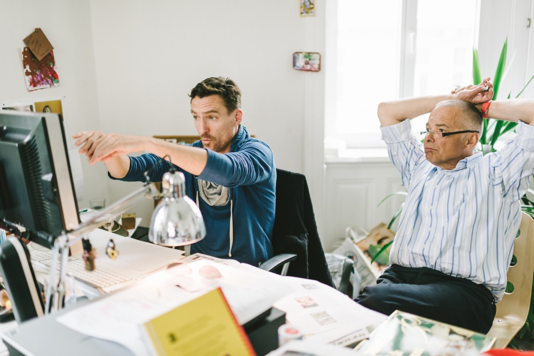 Two men discuss editorial layouts at a desk with computer and design materials in a bright office.