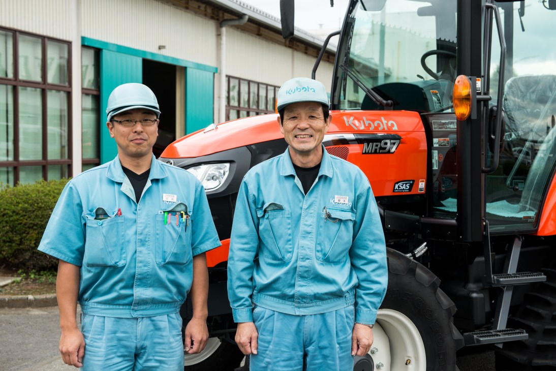 Two Kubota workers in light blue uniforms and white hard hats stand in front of an orange Kubota MR97 tractor at the Tsukuba factory.