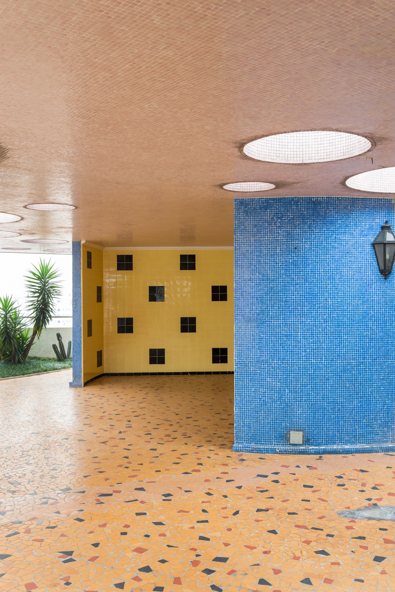 Rooftop area with blue mosaic tile wall, yellow building facade, terracotta floor with geometric pattern, and round skylights in ceiling.