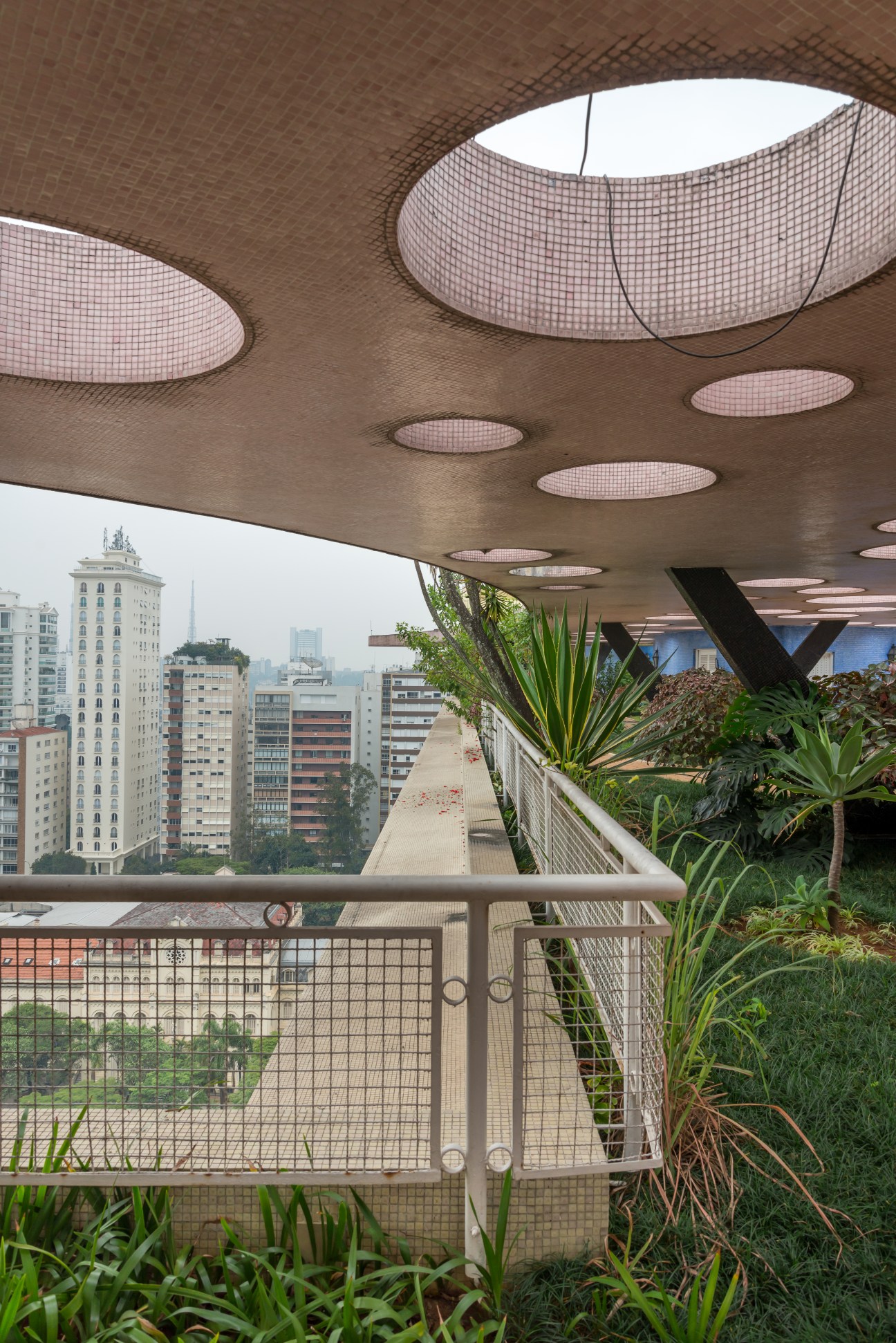 Rooftop covered area with pink mosaic ceiling lights, plants, and São Paulo cityscape view.