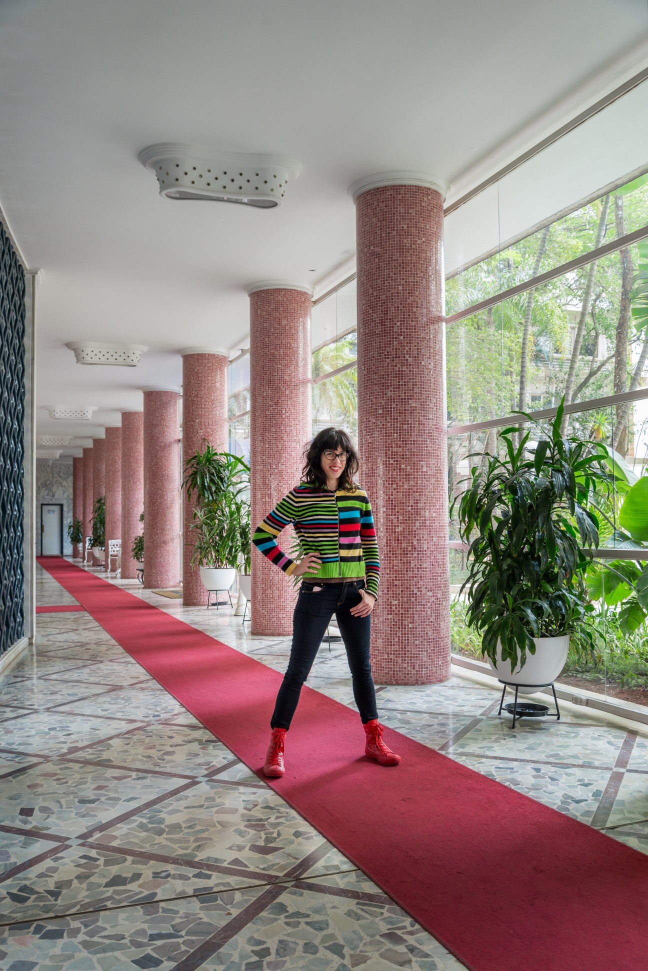 Actress Mariana Eva stands in the Bretagne Building's hallway with red-tiled columns and wine-red carpet.