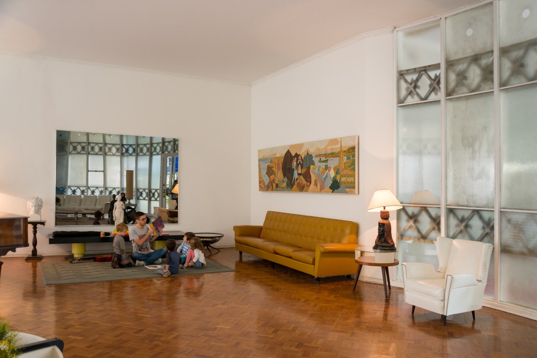 Children sitting on a rug in a modernist communal salon with a mustard sofa, artwork, and large windows.