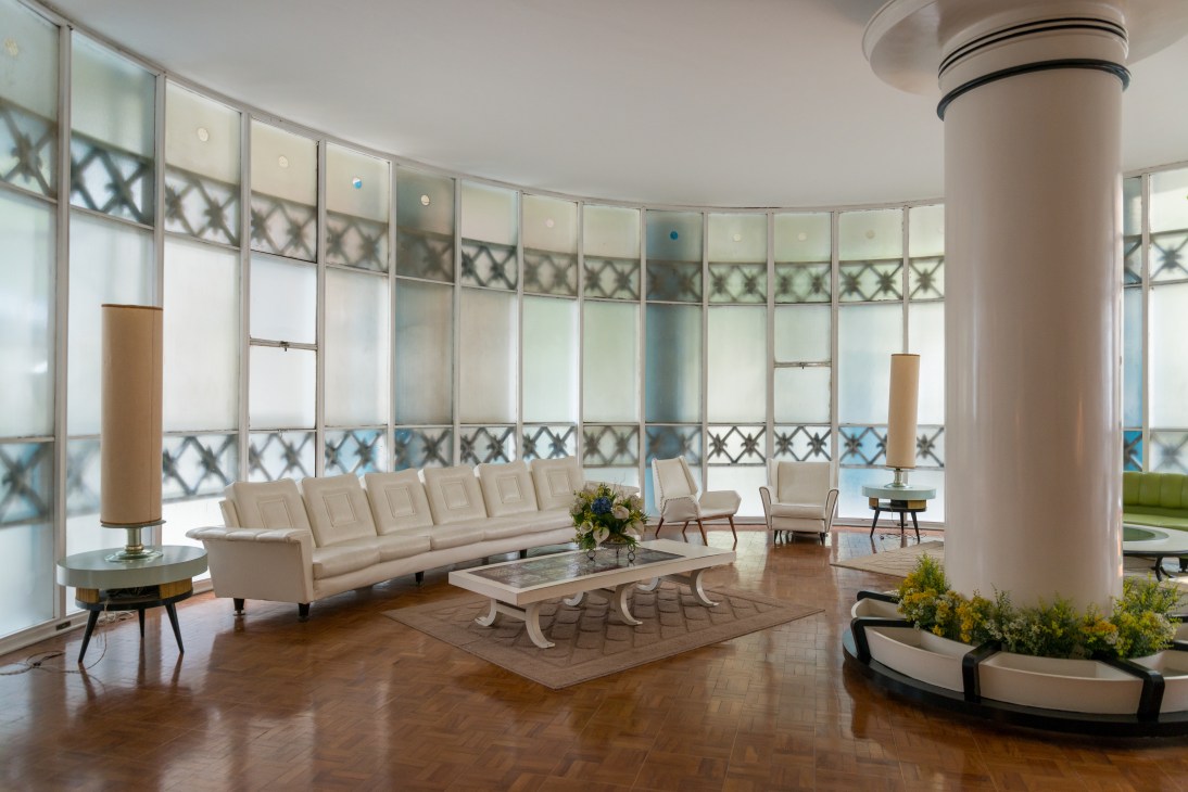 Modern seating area with cream sofa, chairs, and decorative elements in a curved room with floor-to-ceiling windows.