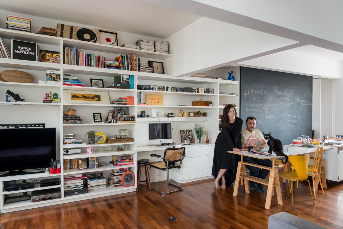 Caroline Baum and Lucio Ribeiro sit at a wooden desk in their modern apartment with white built-in shelving, a black cat nearby.
