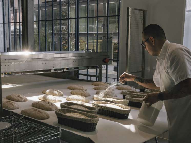 Baker in white shirt dusting flour onto shaped ciabatta loaves at The Manufactory bakery in Los Angeles.