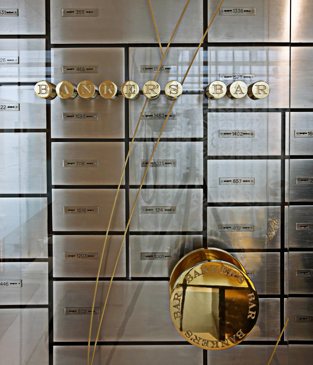 Mandarin Oriental Barcelona's Banker's Bar features brass circular signage spelling 'BANKERS BAR' mounted on a wall of vintage bank deposit boxes with numbered plaques.
