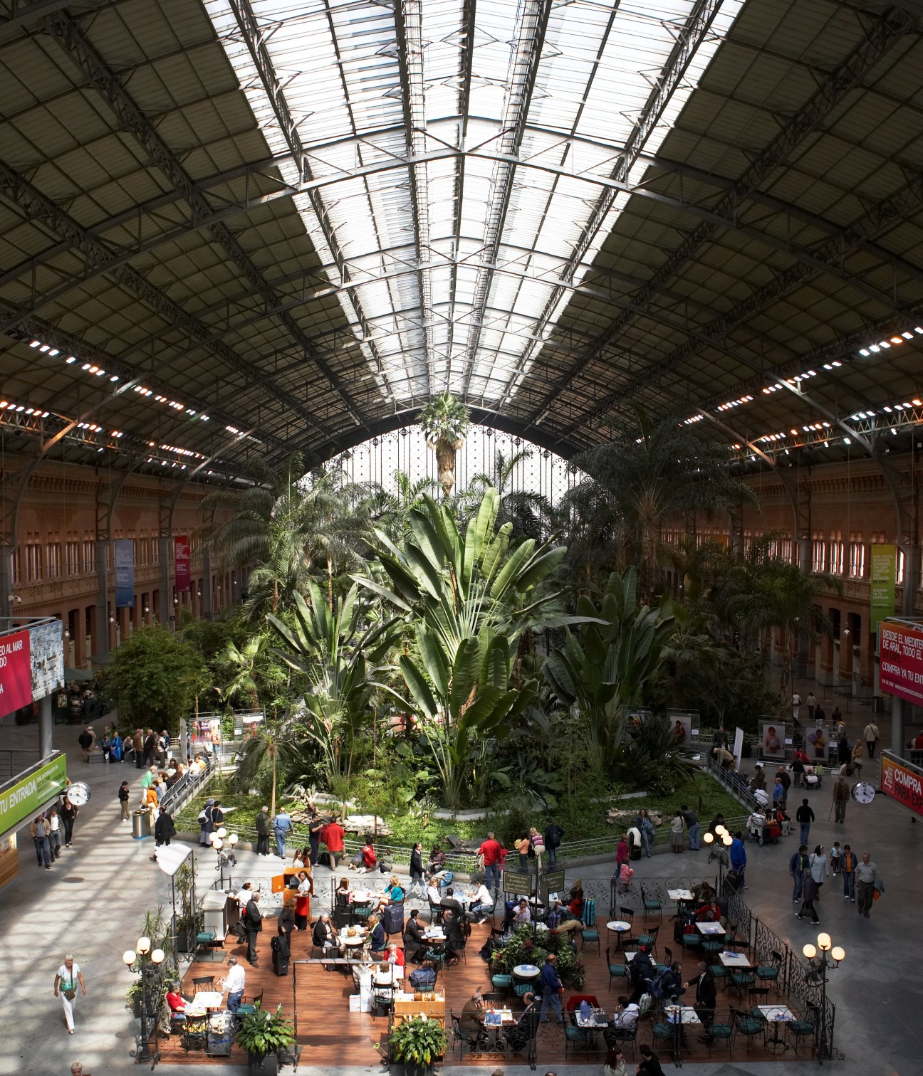 Overhead view of Atocha train station's interior with tropical plants and seated diners under a glass roof.