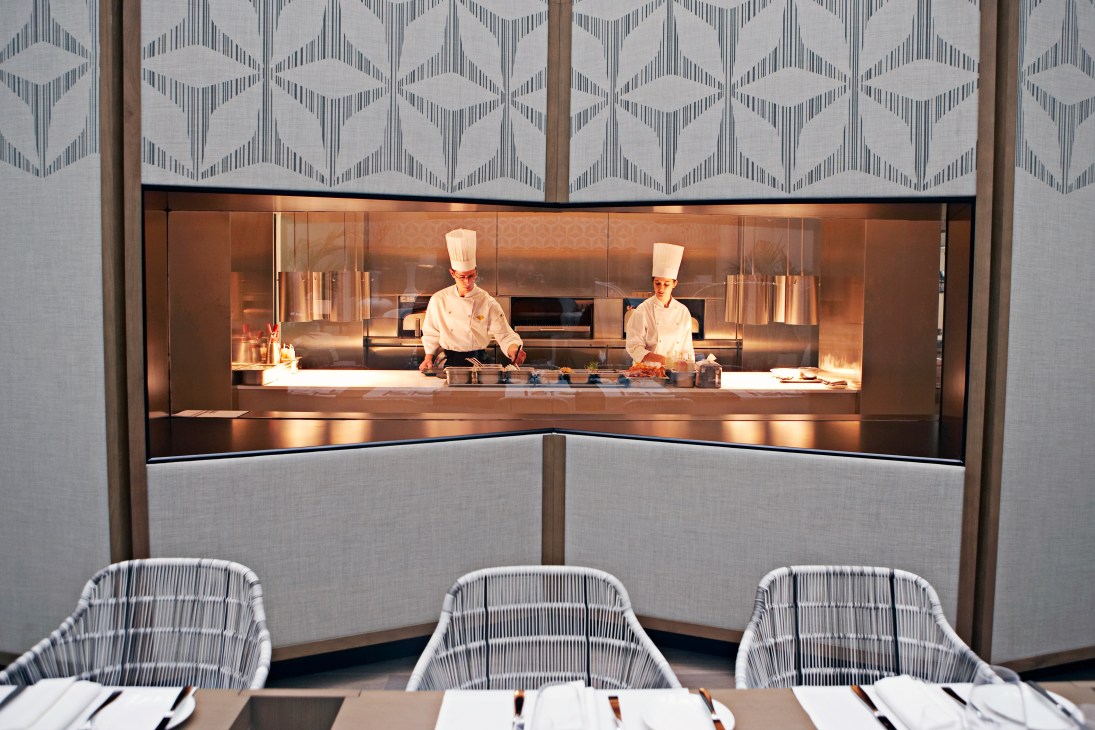 Two chefs prepare food in an open kitchen with copper interior and geometric patterned wall panels above.