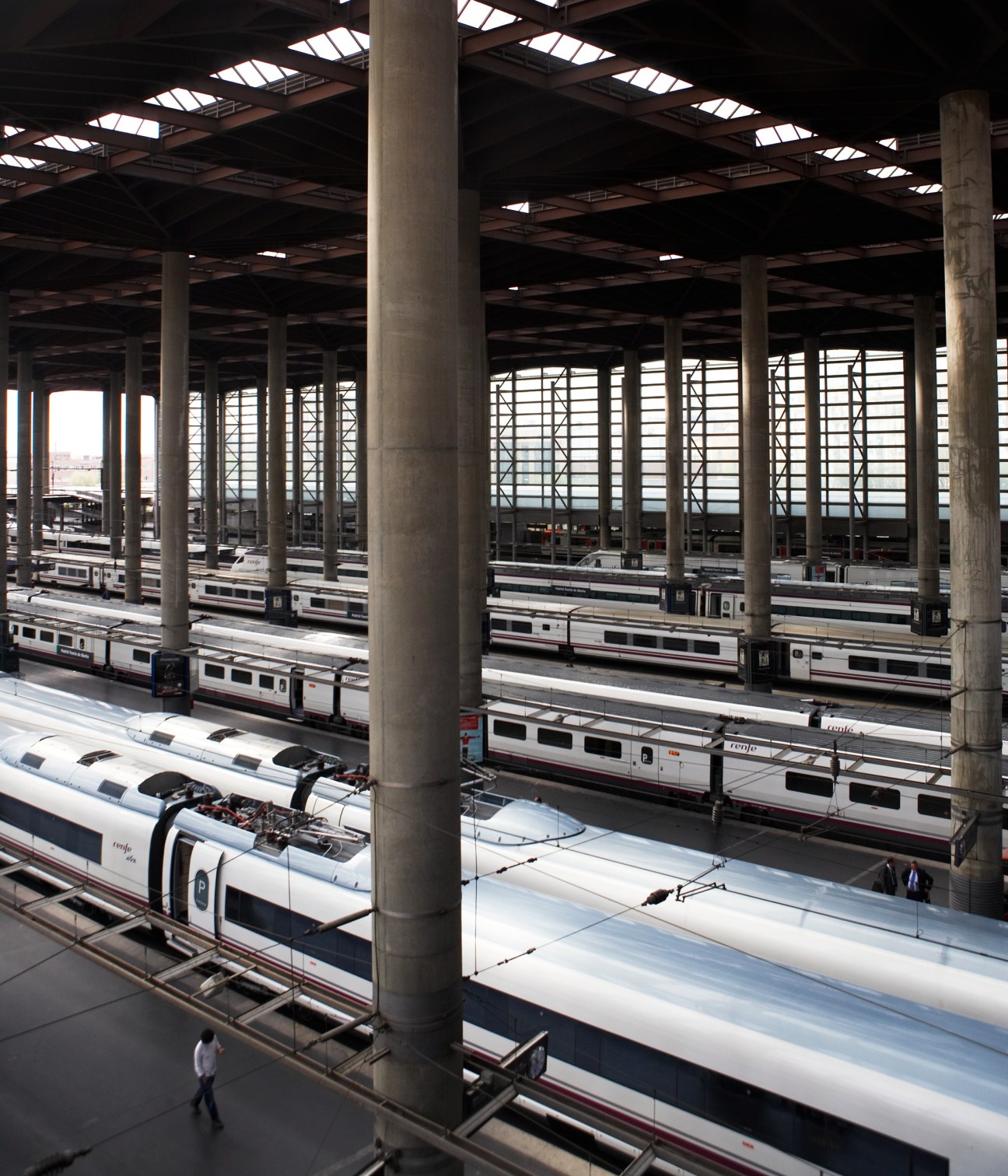 Multiple white and red AVE high-speed trains parked at platforms under a concrete structure at Madrid's Atocha station.
