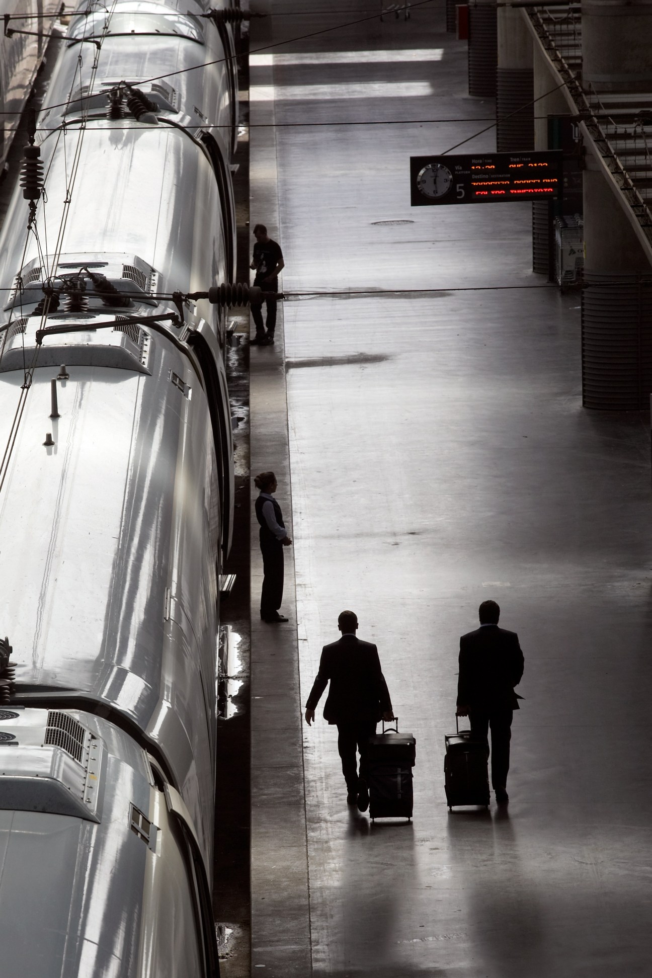 Businesspeople with luggage walking past a silver high-speed train at a station platform with departure board.