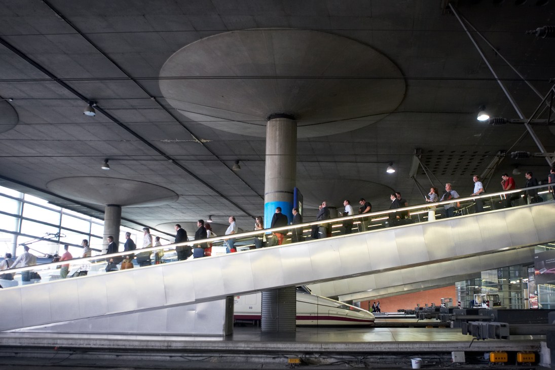 Modern train station platform with sleek white high-speed train and passengers on escalators beneath concrete ceiling.