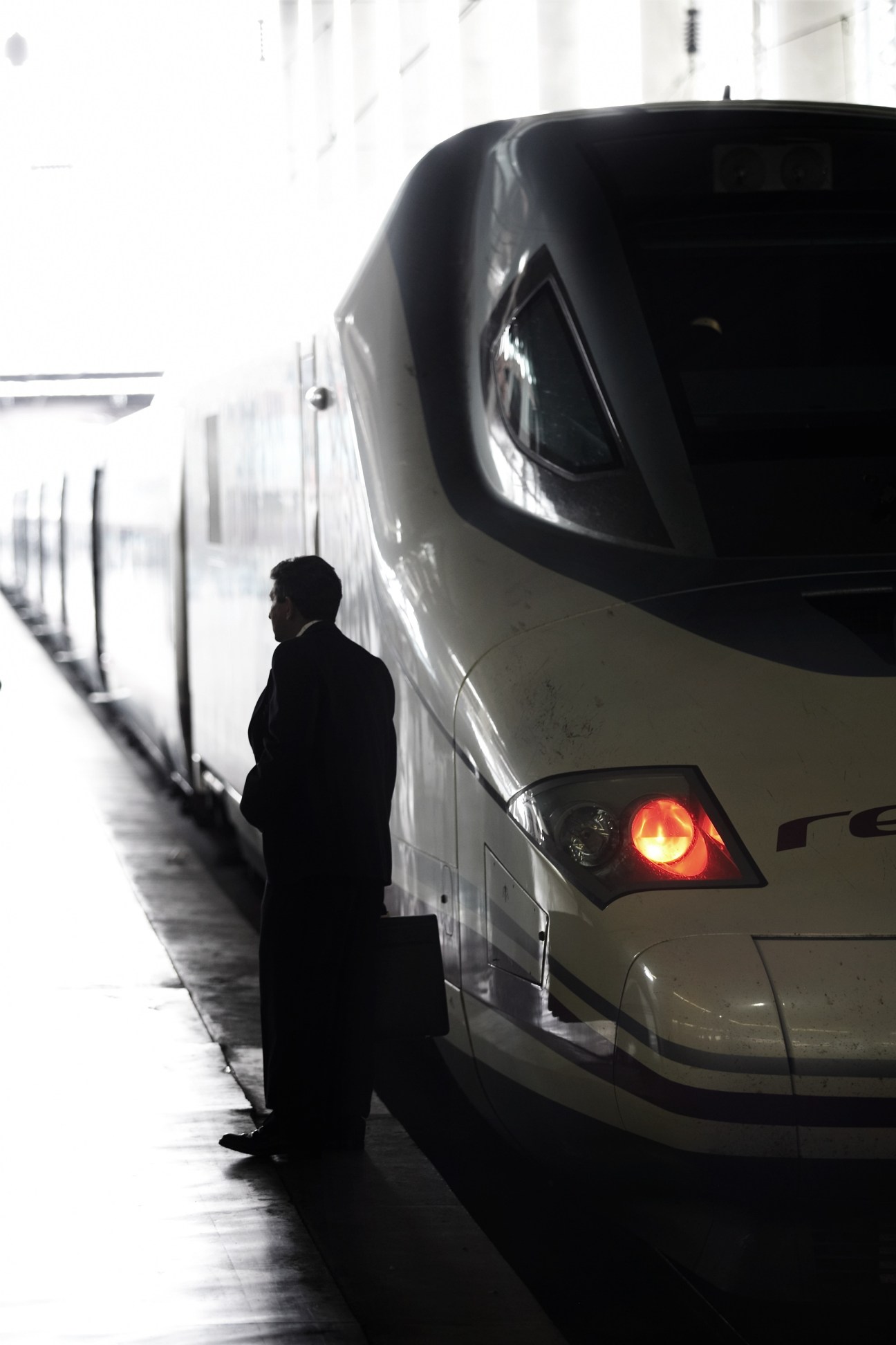 A man in dark clothing stands beside the streamlined nose of a Spanish AVE Series 102 high-speed train at a platform.