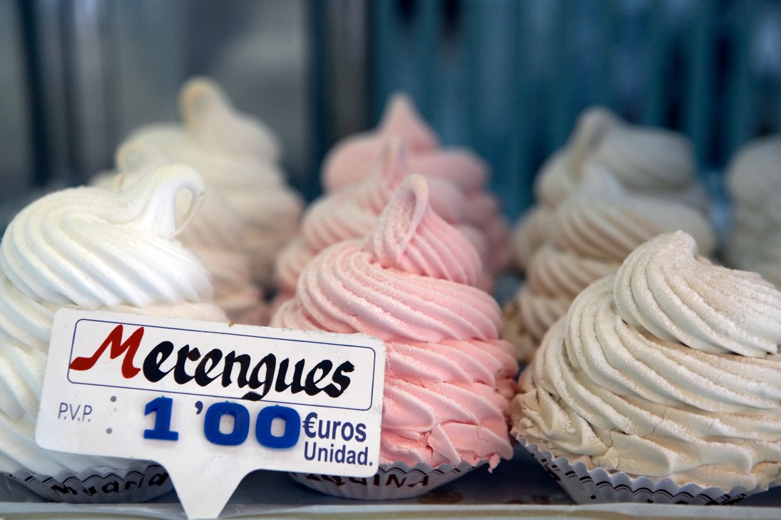 Meringue pastries in white, pink and beige displayed in a bakery window with a price sign reading 1.00€ Unidad.