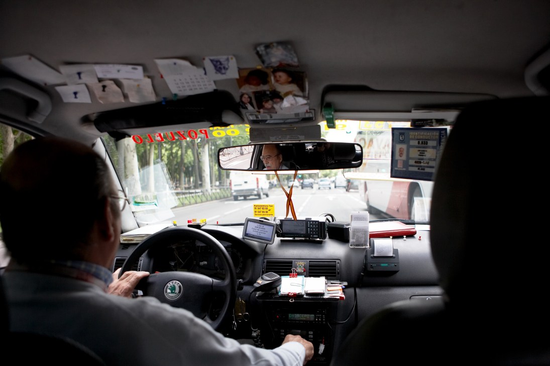 Interior view of a Madrid taxi with driver at wheel, showing cluttered dashboard and personalizations.