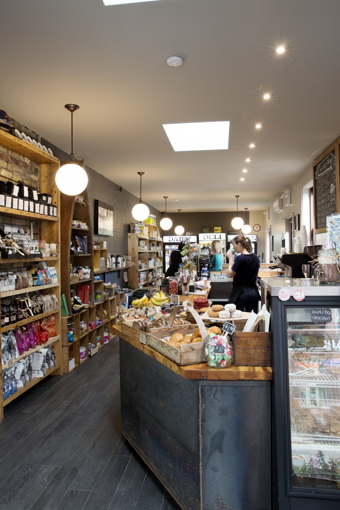 Interior of The Lucky Penny corner shop showing wooden shelves stocked with products, central counter with fresh produce and baked goods, and staff members.