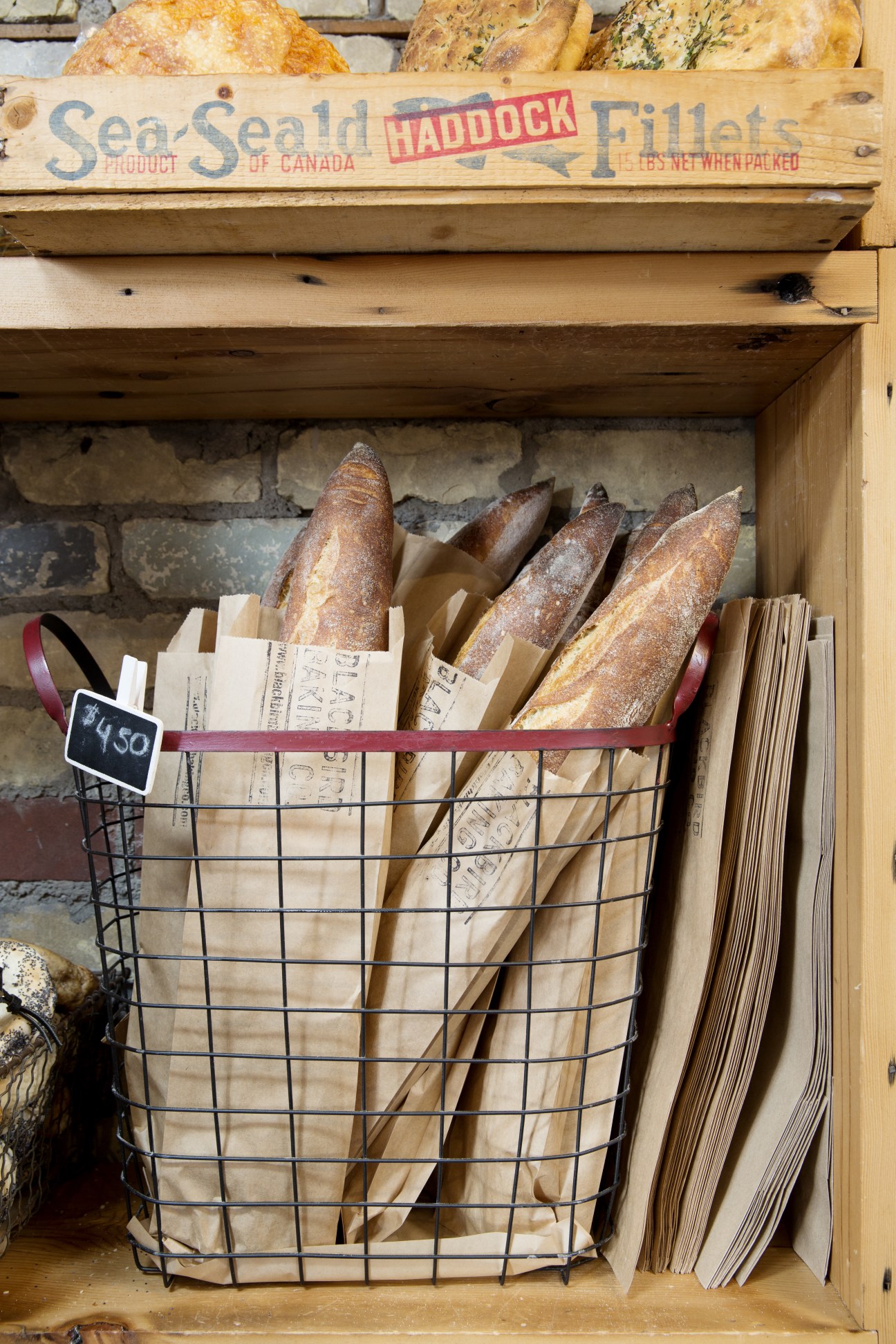 Artisan baguettes wrapped in paper displayed in a wire basket with vintage wooden shelf above.