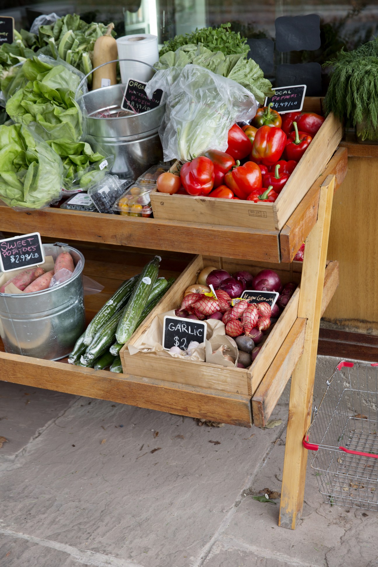 Wooden tiered produce display with fresh lettuce, red peppers, cucumbers, garlic and other vegetables with price signs.