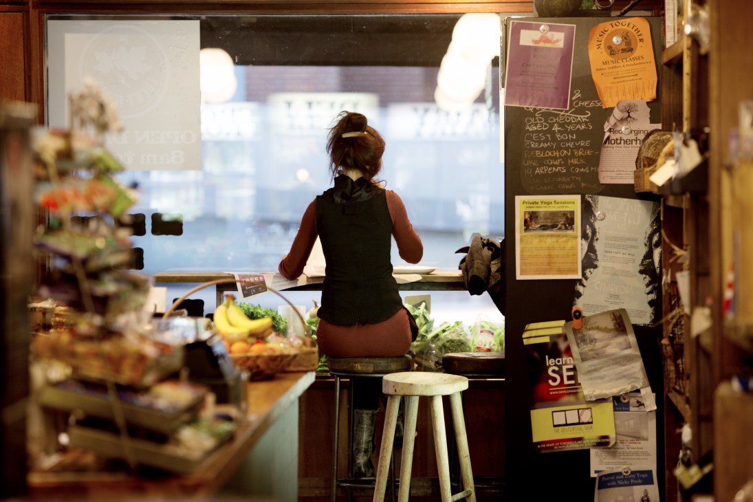 Customer sitting at window seating inside The Lucky Penny corner shop with fresh produce and notices visible.