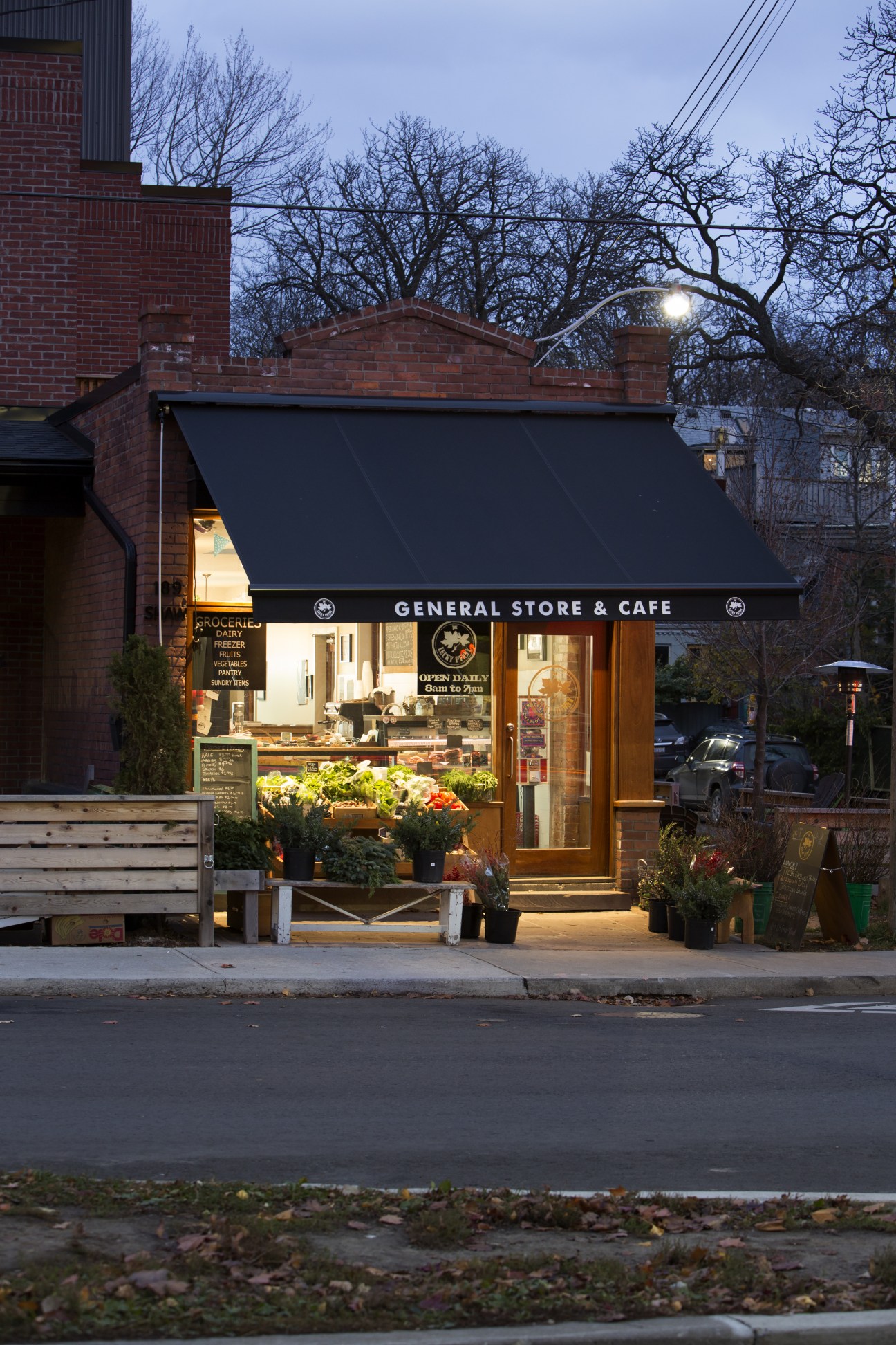 The Lucky Penny general store and café at twilight, with illuminated storefront displaying fresh produce and signage on a Toronto street.