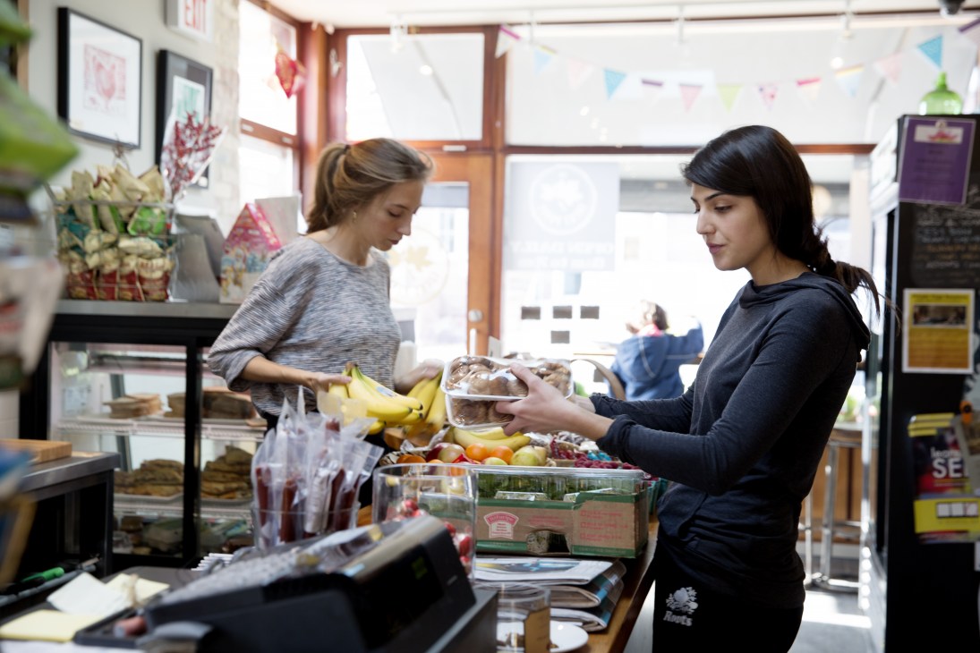 Two cashiers interact with a customer at a corner shop counter displaying fresh produce including bananas.