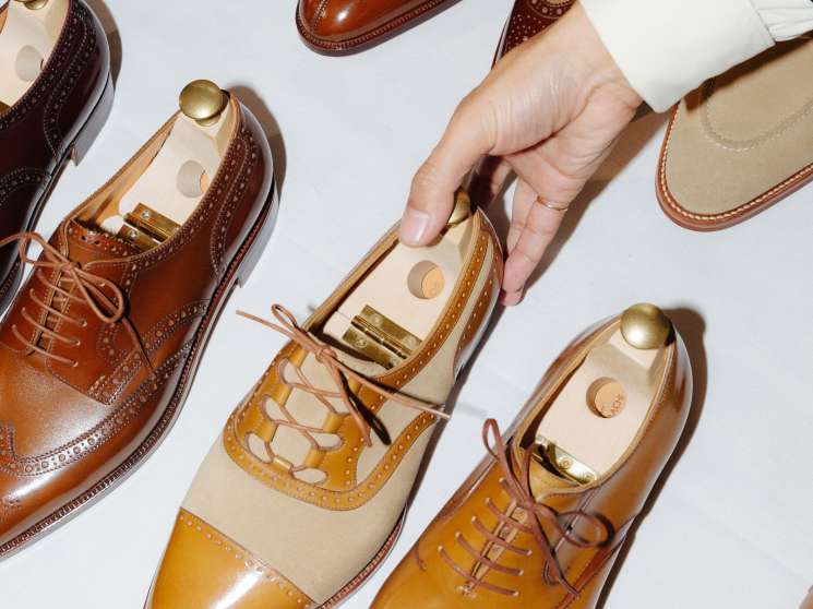 A hand holding a wooden shoe tree inside a polished tan and cream oxford shoe surrounded by other dress shoes.