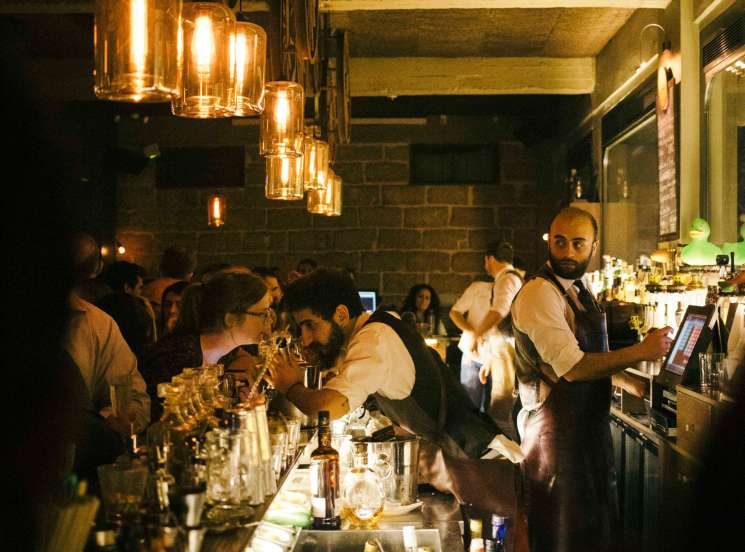 Crowded bar scene with bartender in vest serving drinks under hanging golden lights at night in Beirut.