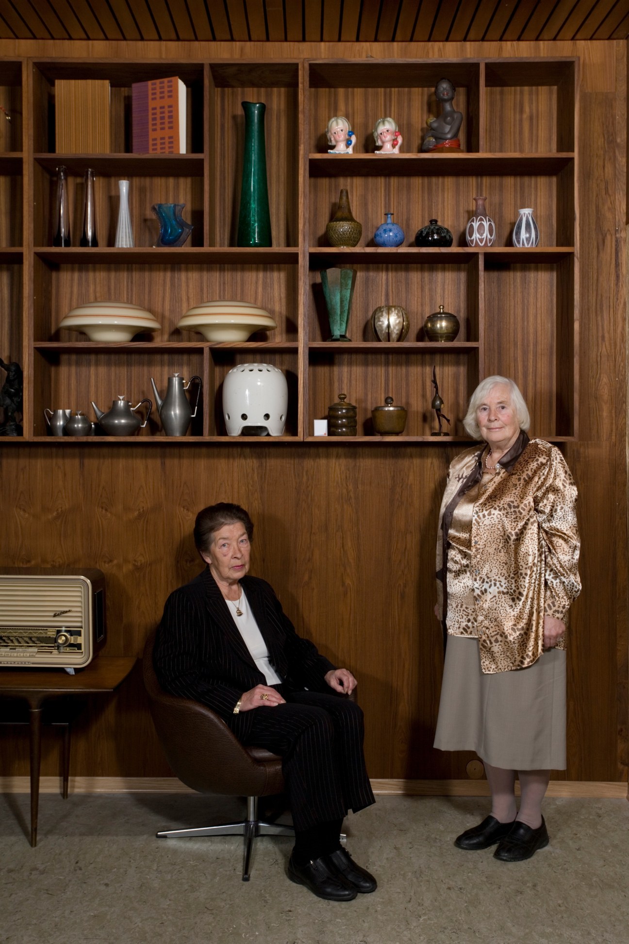 Two elderly women pose in front of wooden shelving displaying Nordic design objects at Fuglen café in Oslo.