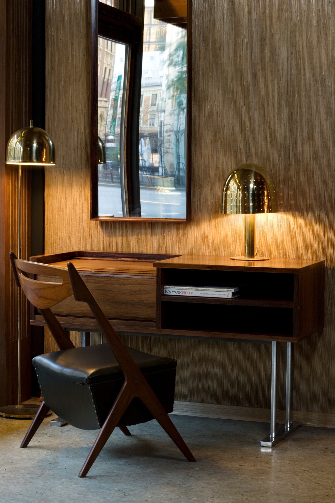 Mid-century modern desk with wooden frame, black chair, and brass dome lamps beside a window with city view.
