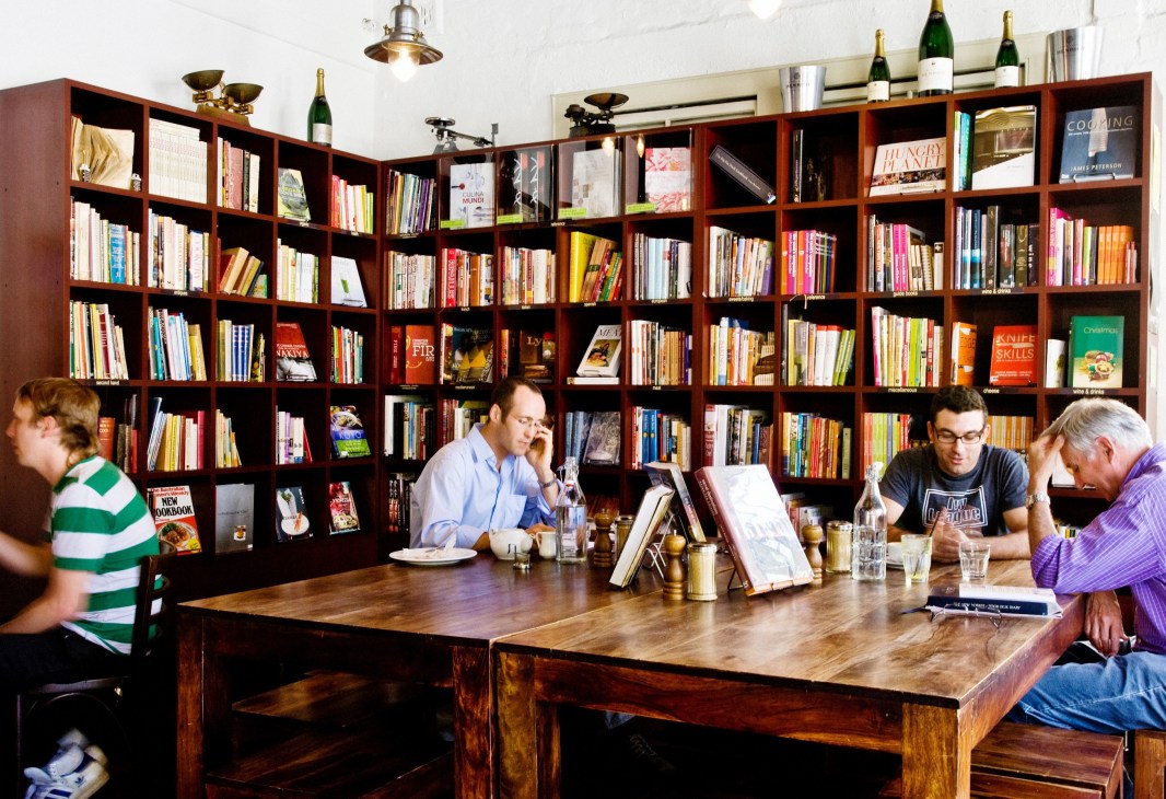 Interior of The Book Kitchen café with customers seated at wooden counter and floor-to-ceiling bookshelves.