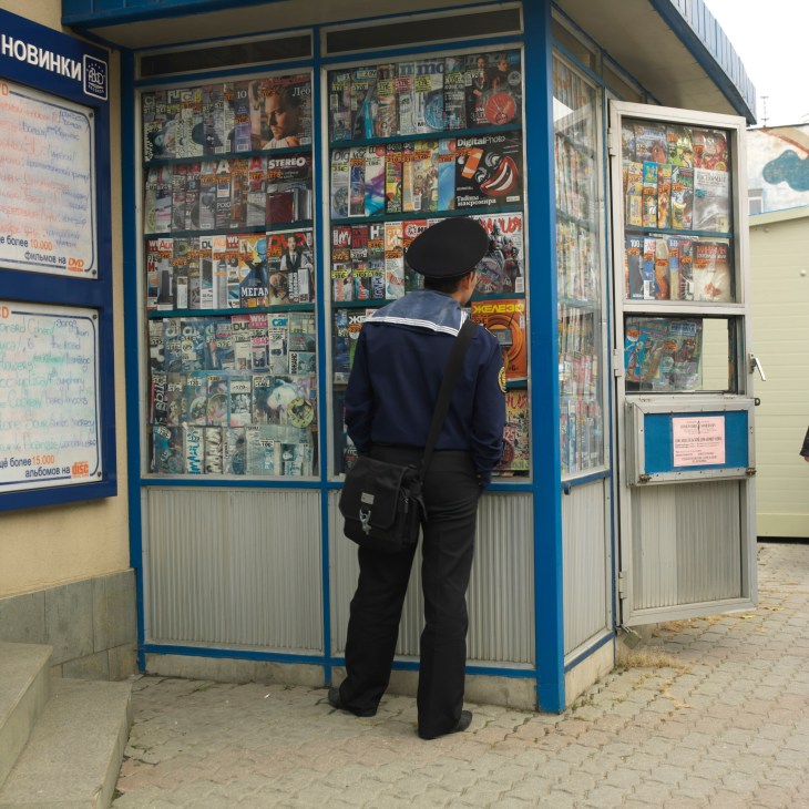 A sailor in navy uniform and black beret browses magazines at a blue kiosk displaying various publications.