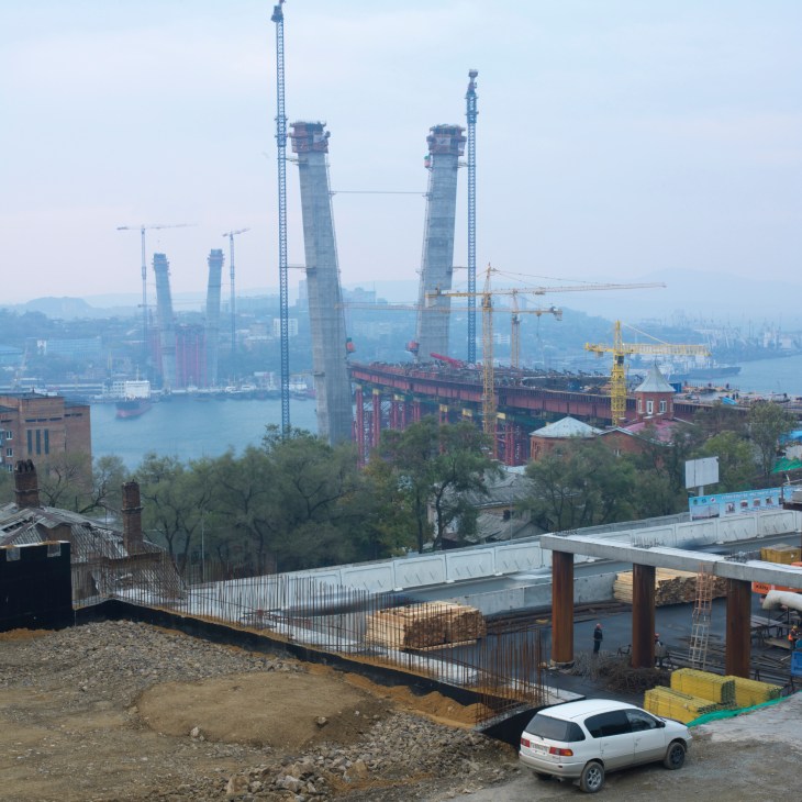 Large bridge under construction with two tall towers spanning Vladivostok harbour, surrounded by construction cranes and equipment.